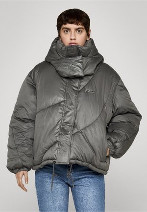 Osoba w szarej, dużej kurtce puchowej z wysokim kołnierzem i niebieskich dżinsach, stojąca na tle jednolitego jasnego tła.