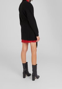 Cárdigan largo negro con cinturón, que presenta un corte holgado y una tela texturizada. Usado sobre un vestido de cuadros rojo y combinado con botas de tacón negras.