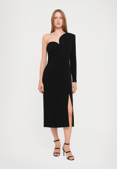 Crna haljina na jedno rame s uskim korzetom, bočnim prorezom i asimetričnim dizajnom. Model nosi minimalne crne sandale s remenčićima i ima elegantnu frizuru.