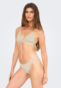 Jonge vrouw met lang bruin haar, gekleed in een beige beha met kant en bijpassende hooggesneden onderbroek, staand tegen een vlakke lichte achtergrond.