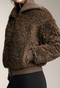 Bruine fleecejas met een gestructureerd, pluizig oppervlak en geribbelde manchetten. Voorzien van een hoge kraag en ritssluiting voor warmte en comfort.