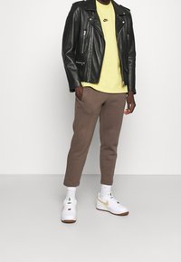 Svart läderbikerjacka, gul Nike-t-shirt, bruna smalrandiga sweatpants, vita sneakers med beige sulor och vita strumpor.