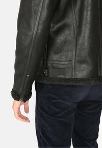 Giacca di pelle nera con finitura testurizzata, polsini in shearling e dettaglio con fibbia nella parte posteriore. Il portatore indossa pantaloni di velluto a coste blu navy.