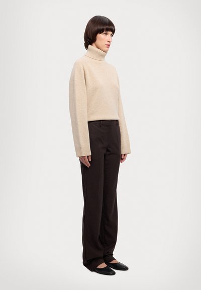 Suéter beige de cuello vuelto con puños acanalados, combinado con pantalones de pierna ancha marrón oscuro y zapatos planos negros, presentado sobre un fondo liso.