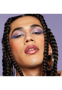 Nahaufnahme eines Gesichts mit geflochtenem Haar, das große transparente Ohrringe trägt. Lila Lidschatten und glänzende Lippen auf glatter Haut. Sanfte Beleuchtung.