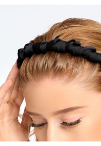 Femme aux cheveux châtain clair portant un bandeau en tissu noir froncé, touchant le côté de sa tête avec la main, les yeux fermés.