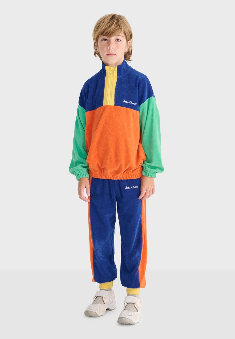 Kind trägt einen farblich abgesetzten Trainingsanzug mit blauen, grünen, orangefarbenen und gelben Abschnitten und steht vor einem schlichten hellen Hintergrund.