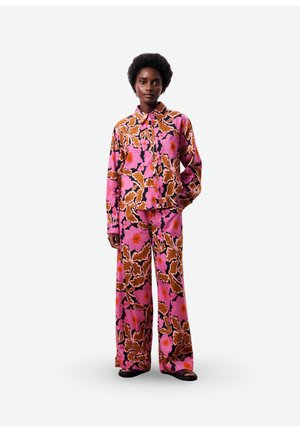 Femme debout portant une chemise à manches longues à motif floral rose assortie et un pantalon large avec des sandales noires sur fond gris.