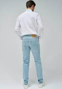 Jeans de mezclilla azul claro con corte ajustado, diseño de cinco bolsillos y una etiqueta de cuero marrón en la parte posterior. Llevados con una camisa blanca de manga larga.