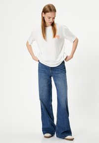 Hvid bomulds T-shirt med orange "California 98" tryk, parret med flagrende blå denim jeans. Modellen står med hænderne på hoften og ser ned.