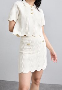 Ensemble blanc deux pièces composé d'un haut court à ourlet festonné avec des boutons dorés et d'une jupe assortie ornée de détails décoratifs à boutons et de texture.