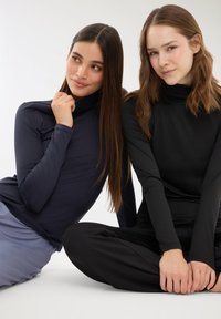 Dwie kobiety siedzą na podłodze, ubrane w długie rękawy golfy; jedna w granatowej bluzce i fioletowych spodniach, a druga w czarnej bluzce i pasujących spodniach.