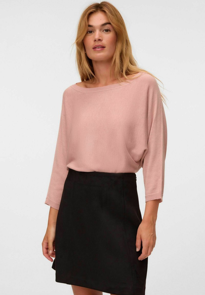 Femme aux cheveux blonds portant un haut rose ample à manches longues rentré dans une jupe noire simple longueur genou, sur un fond uni.