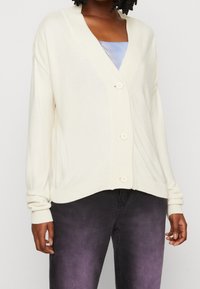 Krämfärgad kofta med V-ringning, med ribbat mönster och tre stora knappar, kombinerad med mörka jeans med blekningar.