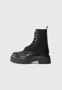 Black lace-up leather boot with thick rubber sole and knitted stretchy ankle panel, side view on plain light background.