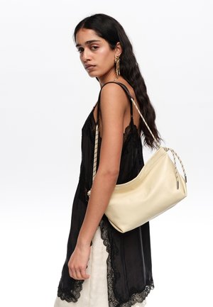 Pikkade tumedate juustega naine, kandes musta pitsiga ääristatud kleiti, kandmas kreemjat nahast õlakotti köisrihmadega.