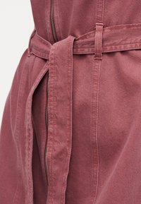 Κοντινό πλάνο ρουχισμού σε σκόπιμο ροζ χρώμα με υφασμάτινη ζώνη δεμένη γύρω από τη μέση και εμφανές κάθετο φερμουάρ στο κέντρο.