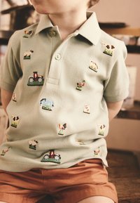 Polo vert clair avec des animaux de ferme et des tracteurs brodés, doté d'un col et de manches courtes. Associé à un short marron.