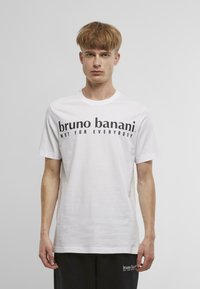 Bílý bavlněný tričko s černým textem "bruno banani. NENÍ PRO KAŽDÉHO." Kulatý výstřih, krátké rukávy a rovný střih.