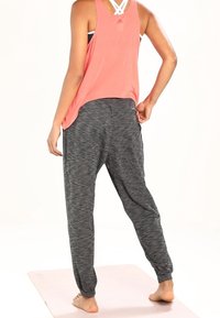 Frau, die barfuß auf einer Yogamatte steht, einen korallfarbenen Tanktop und graue Jogginghosen trägt, mit dem Rücken zu uns und einer Hand auf der Hüfte.