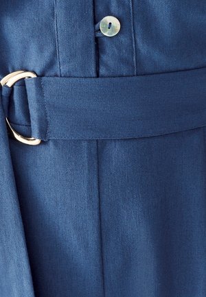 Blauer Stoffgürtel, der durch eine silberne D-Ring-Schnalle auf einem Kleidungsstück gefädelt ist, mit einem sichtbareren Knopf aus Perlmutt oben.