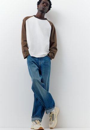 En ung mand læner sig op ad en hvid væg med hænderne i lommerne, iført en brunt og hvid langærmet skjorte, blå jeans og beige sneakers.