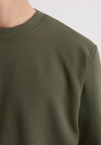 T-shirt à manches longues vert olive en coton doux, avec un col ras du cou et une texture lisse. Coutures visibles le long du col.