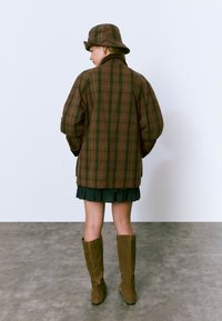 Persona con chaqueta de cuadros marrón y sombrero de cubo a juego, falda corta negra y botas marrones hasta la rodilla, de pie sobre un suelo gris.