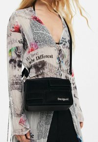 Kobieta nosząca przezroczystą bluzkę z tekstem i kwiatowymi nadrukami, trzymająca czarną torebkę crossbody Desigual z detalami patchworkowymi.