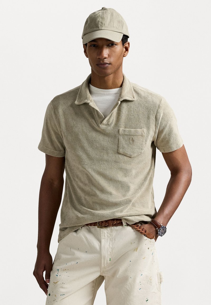 Polo shirt en velours de couleur kaki avec un col et une poche poitrine, associé à un pantalon crème présentant des éclaboussures de peinture et une ceinture tressée marron.