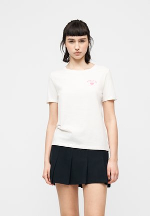 Jeune femme aux cheveux foncés portant un t-shirt blanc à manches courtes avec un texte rose et une mini-jupe plissée noire sur un fond uni.