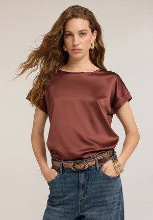Kvinde med langt bølget hår iført gyldne hoop-øreringe, mørkerød satinbluse, blå jeans med brun bælte og gyldne armbånd med hænderne i lommerne.