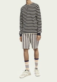 Gestreiftes Langarmshirt in Marineblau und Cremé, kombiniert mit vertikal gestreiften Shorts in Beige und Braun. Creméfarbene Sneaker und mehrfarbige Socken.
