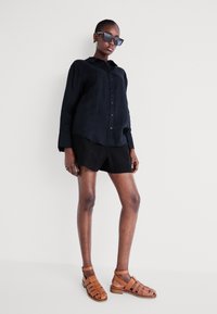 Schwarzes Leinenhemd mit Knöpfen, kombiniert mit schwarzen Shorts und braunen Riemensandalen. Zubehör umfasst große eckige Sonnenbrillen.