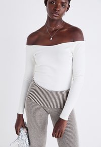 Off-shoulder, långärmad vit ribbad topp i kombination med tajta, texturerade byxor i beige och bruna ränder. Modellen håller en silverglittrande clutch.