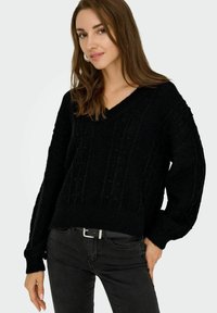 Pull en maille noir avec un col en V, motif texturé et coupe décontractée. Manches longues et poignets côtelés. Porté avec un jean foncé et une ceinture noire.