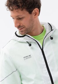 Giacca leggera e traslucida color verde menta con dettagli neri, cappuccio regolabile e zip laterale. Presenta un testo stampato: "L'UNICO LIMITE SEI TU."