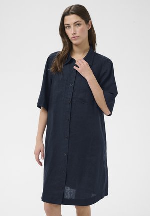 Femme aux longs cheveux bruns portant une robe-chemise ample, boutonnée, de couleur bleu marine foncé avec des manches courtes, sur un fond uni.