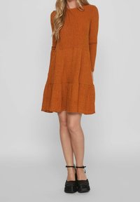 Abito a coste di colore ruggine con maniche lunghe e design a strati, caratterizzato da un colletto rotondo e una vestibilità ampia. Le scarpe con plateau nere completano il look.
