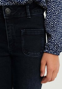 Mörkblå denimjeans som har en bakficka, kompletterade med en långärmad skjorta med ett vitt prickigt mönster på en marinblå bakgrund.