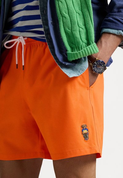 Oransje svømmeshorts med snorsøm, med et brodert bjørnmønster på venstre side og glatt, lett stoff.