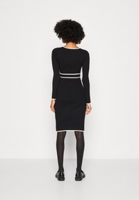 Robe noire en maille côtelée avec manches longues, accents blancs au niveau du col et de la taille, silhouette ajustée, associée à des collants et des chaussures noirs.