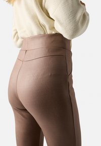 Pantalones marrones de cintura alta hechos de una tela lisa y elástica. Presentan un diseño que contornea la figura con costuras sutiles a lo largo de la parte trasera.