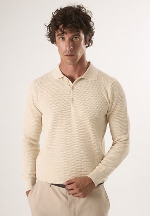 Beige gestricktes Polohemd mit langen Ärmeln, das ein strukturiertes Muster, einen klassischen Kragen und eine Knopfleiste mit drei Knöpfen aufweist. Getragen mit beigen Hosen.