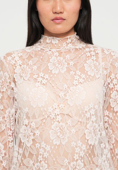 Doorzichtig kant top met bloemenpatroon, hoge halslijn, lange mouwen, in zachte beige tinten. De textuur is delicaat en transparant, wat de elegantie versterkt.