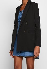 Blazer negro con un diseño ajustado, dos bolsillos frontales y detalles en tono dorado. Llevado sobre un vestido de mezclilla, con una forma estructurada.