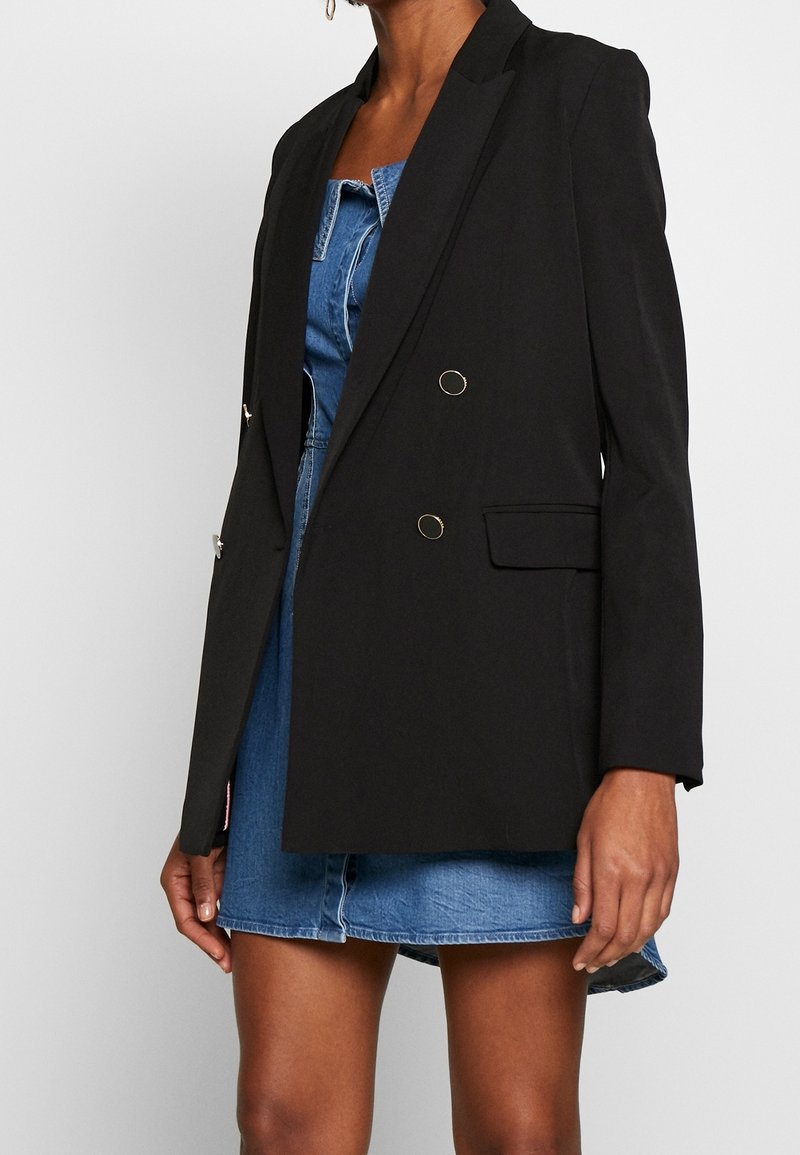 Blazer negro con un diseño ajustado, dos bolsillos frontales y detalles en tono dorado. Llevado sobre un vestido de mezclilla, con una forma estructurada.