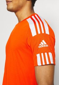 Světlý oranžový sportovní tričko s bílými proužky na rukávech. Na levém rameni je malá bílá značka Adidas.