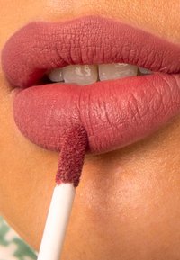 Matte mauve leppestift påført med en flat applikator på fyldige lepper, som viser en jevn tekstur mot en lett opplyst hudtone.