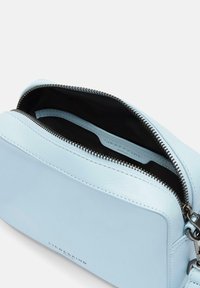 Jasnoniebieska skórzana torba z zapięciem na zamek, z zamszowym wnętrzem i małą wewnętrzną kieszonką. Widoczna nazwa marki wytłoczona z przodu.